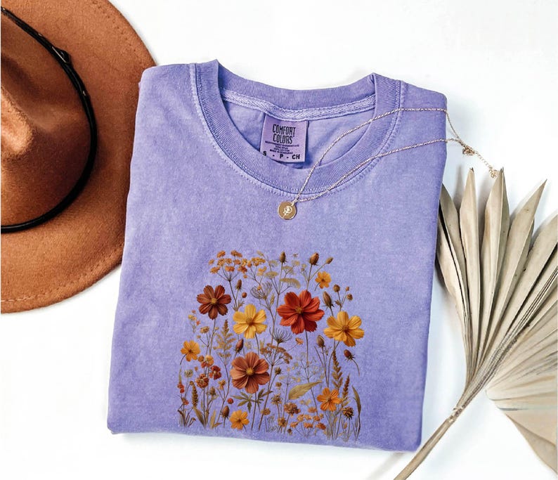 K&ouml;nnte beinhalten: Ein lavendelfarbenes Kurzarm-T-Shirt mit einem Comfort Colors-Etikett am Halsausschnitt. Das Shirt zeigt einen Grafikdruck verschiedener Wildblumen in Orange-, Gelb- und Braunt&ouml;nen. Eine goldfarbene Halskette mit einem kleinen runden Anh&auml;nger liegt auf dem Stoff.
