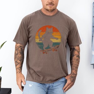May include: A brown t-shirt with a retro-style graphic of a cat skateboarding. The cat is gray and the graphic is set against a sunset background with orange, yellow, and red colors.