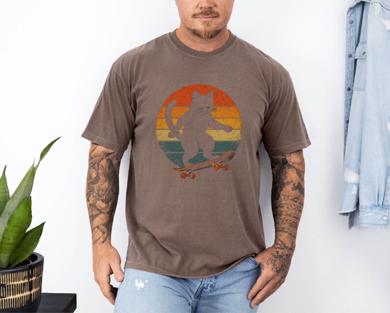 May include: A brown t-shirt with a retro-style graphic of a cat skateboarding. The cat is gray and the graphic is set against a sunset background with orange, yellow, and red colors.