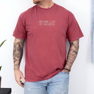 May include: A maroon-colored t-shirt with the text "AWESOME LIKE MY DAUGHTER" in white lettering. The shirt is a crew neck style and appears to be made of a soft, comfortable material. The shirt is worn with blue jeans.