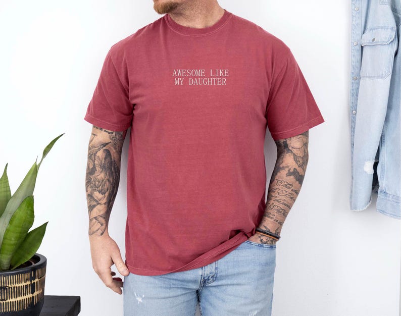 May include: A maroon-colored t-shirt with the text "AWESOME LIKE MY DAUGHTER" in white lettering. The shirt is a crew neck style and appears to be made of a soft, comfortable material. The shirt is worn with blue jeans.