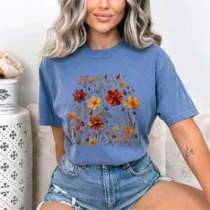 K&ouml;nnte beinhalten: Ein blaues kurz&auml;rmeliges T-Shirt mit Rundhalsausschnitt und einem zentralen Grafikdruck verschiedener Wildblumen in Orange-, Gelb- und Dunkelrott&ouml;nen. Das Design zeigt eine Ansammlung von St&auml;ngeln, Bl&auml;ttern und bl&uuml;henden Blumen.