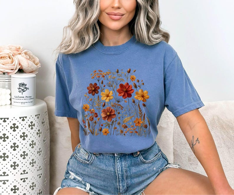 K&ouml;nnte beinhalten: Ein blaues kurz&auml;rmeliges T-Shirt mit Rundhalsausschnitt und einem zentralen Grafikdruck verschiedener Wildblumen in Orange-, Gelb- und Dunkelrott&ouml;nen. Das Design zeigt eine Ansammlung von St&auml;ngeln, Bl&auml;ttern und bl&uuml;henden Blumen.