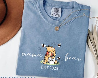 May include: Blue jean Comfort Colors t-shirt with a Winnie the Pooh graphic. The graphic includes the words "mama bear" and "bear", Winnie the Pooh, bees, and the year "EST.2025". A gold necklace is draped over the shirt.