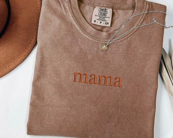 May include: A brown short-sleeved t-shirt with the word mama embroidered in orange thread across the chest. The shirt features a Comfort Colors label at the inner neckline and is accompanied by a silver-toned necklace with a small circular pendant.