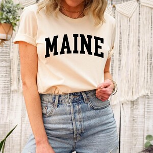 May include: A cream-colored short-sleeve t-shirt featuring the word MAINE printed in large, bold, black capital letters across the chest in an arched collegiate font, paired with light blue denim jeans with a visible metal button and rivets.
