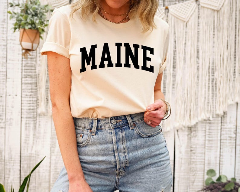 May include: A cream-colored short-sleeve t-shirt featuring the word MAINE printed in large, bold, black capital letters across the chest in an arched collegiate font, paired with light blue denim jeans with a visible metal button and rivets.