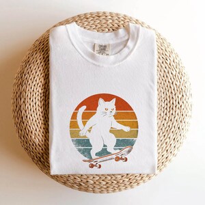 May include: White cat skateboarding on a vintage sunset background. The cat is wearing a white t-shirt with a red and blue logo.