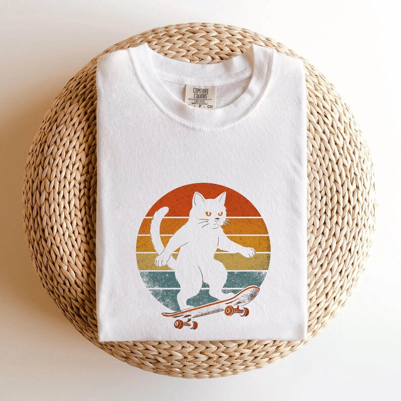 May include: White cat skateboarding on a vintage sunset background. The cat is wearing a white t-shirt with a red and blue logo.