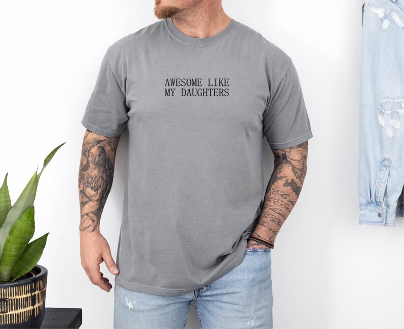 May include: A gray t-shirt with the text "AWESOME LIKE MY DAUGHTERS" in black. The shirt is worn by a person with tattoos on their arms and is paired with blue jeans. A denim jacket hangs in the background.