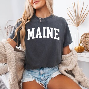 May include: A charcoal gray short-sleeve t-shirt featuring the word MAINE printed in large white capital letters across the chest in a classic arched collegiate font. The shirt has a crew neckline and a relaxed fit with a vintage-style wash.