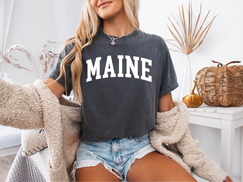May include: A charcoal gray short-sleeve t-shirt featuring the word MAINE printed in large white capital letters across the chest in a classic arched collegiate font. The shirt has a crew neckline and a relaxed fit with a vintage-style wash.