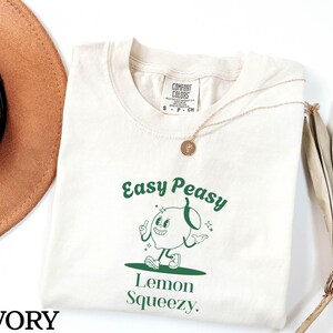 May include: Ivory t-shirt with a green graphic of a cartoon lemon with the text "Easy Peasy Lemon Squeezy."