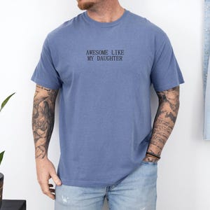 May include: A blue short-sleeve t-shirt with the text "AWESOME LIKE MY DAUGHTER" in black. The shirt is worn by a person with tattoos on their arms. A denim jacket hangs on the wall in the background.