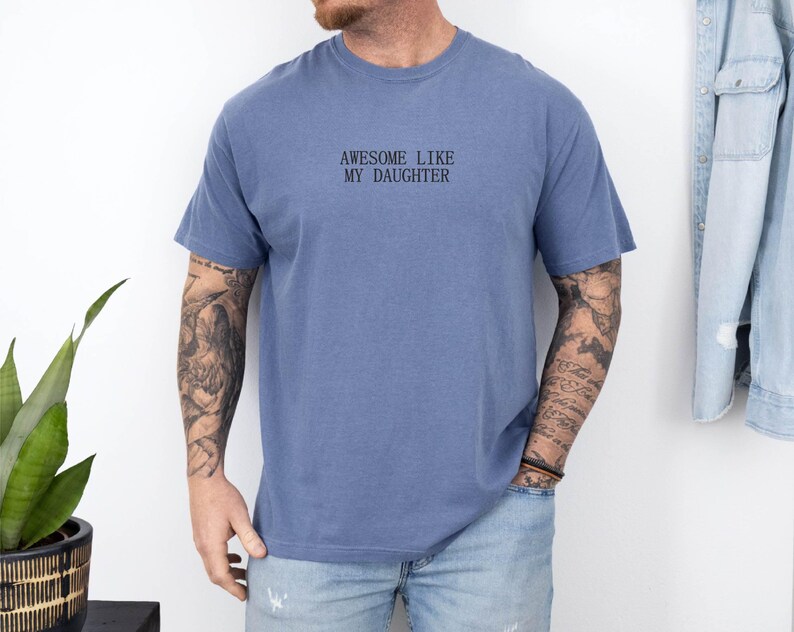 May include: A blue short-sleeve t-shirt with the text "AWESOME LIKE MY DAUGHTER" in black. The shirt is worn by a person with tattoos on their arms. A denim jacket hangs on the wall in the background.