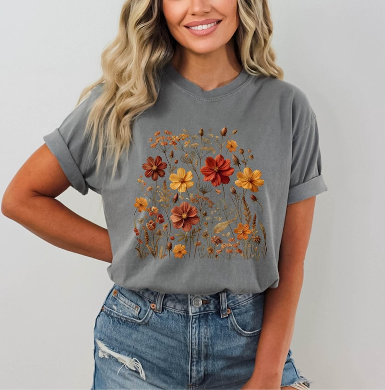 K&ouml;nnte beinhalten: Ein anthrazitfarbenes Kurzarm-T-Shirt mit einem zentralen Grafikdruck verschiedener Wildblumen in Orange-, Gelb- und Rostt&ouml;nen. Das Design zeigt detaillierte Illustrationen von bl&uuml;henden Blumen, Knospen und d&uuml;nnen St&auml;ngeln in einer Anordnung.