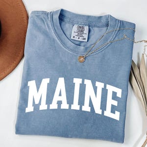 May include: A blue short-sleeved t-shirt with the word MAINE printed in large white capital letters across the chest. The shirt features a crew neckline and a Comfort Colors brand label on the inside collar indicating a size small.