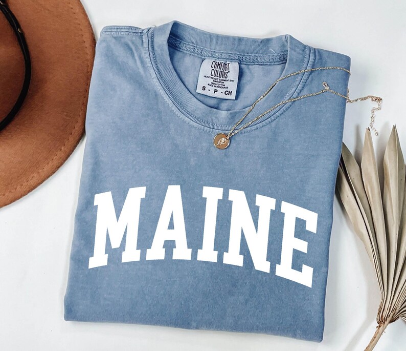 May include: A blue short-sleeved t-shirt with the word MAINE printed in large white capital letters across the chest. The shirt features a crew neckline and a Comfort Colors brand label on the inside collar indicating a size small.