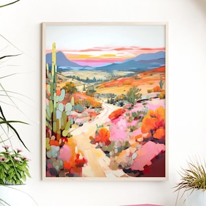 May include: A framed watercolor painting depicts a desert landscape with a winding dirt road. The artwork features vibrant colors, including pink, orange, and green, with a sunset in the background. The painting is displayed on a white shelf with plants.