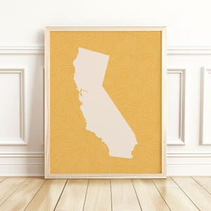 May include: A framed print featuring a cream-colored outline of California against a yellow background with a topographic pattern. The print is displayed on a wooden floor against a white wall.