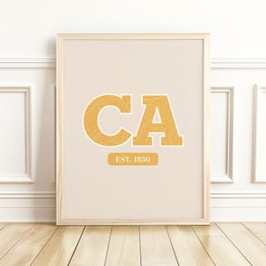 May include: A framed print featuring the letters "CA" in a bold, yellow font with a topographic design. Below the letters is a yellow rectangle with the text "EST. 1850". The background is a light beige color. The frame is a light wood color.