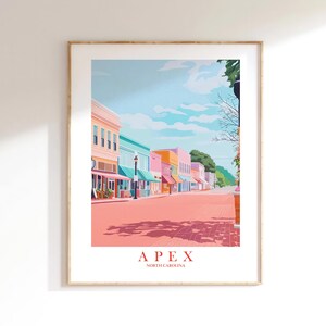 May include: A framed art print showing a street scene in Apex, North Carolina. The image displays a row of buildings in pastel shades, a pink road, and a blue sky with clouds. The words "APEX" and "NORTH CAROLINA" are printed at the bottom of the artwork.