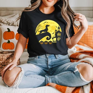 May include: Black t-shirt featuring a Halloween-themed design. The graphic includes a full yellow moon, a silhouette of a dachshund dog, a haunted house, bats, and jack-o'-lanterns. The shirt is made of a soft material and is suitable for casual wear.