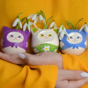 May include: Three ceramic cat-shaped vases in purple, white, and blue, each holding white snowdrop flowers. The vases have cat faces with closed eyes and a bow. The white vase has a green floral pattern. The background is yellow.
