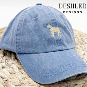 May include: A light blue denim baseball cap with an embroidered image of a golden retriever dog wearing a blue collar. The cap has a curved brim and a mesh back. The text "DESHLER DESIGNS" is printed in white on the back of the cap.