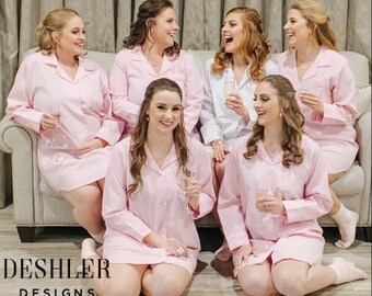 May include: Six people wearing pink pajama sets are gathered together, some sitting on a light-colored couch and others kneeling on the floor. The pajama sets have a button-down shirt style. The text "DESHLER DESIGNS" is visible at the bottom of the image.