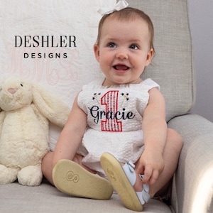 May include: A baby girl wearing a white sleeveless dress with a red and white striped number one and the name "Gracie" embroidered on it. The dress has a red, white, and blue firework design. The baby is sitting on a chair and smiling.