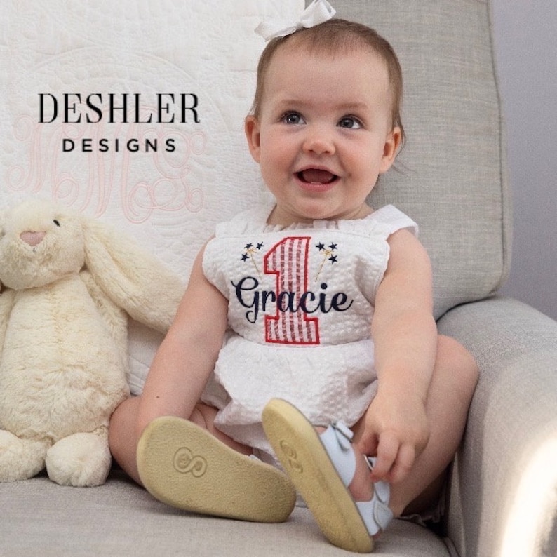 May include: A baby girl wearing a white sleeveless dress with a red and white striped number one and the name "Gracie" embroidered on it. The dress has a red, white, and blue firework design. The baby is sitting on a chair and smiling.