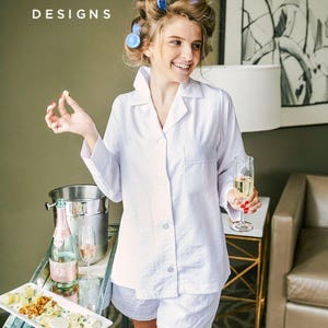 Puede incluir: Una mujer con una camisa de pijama blanca con botones y pantalones cortos a juego. Est&aacute; sosteniendo una copa de champ&aacute;n y un peque&ntilde;o trozo de comida. El texto "DESHLER DESIGNS" es visible en la esquina superior izquierda de la imagen.