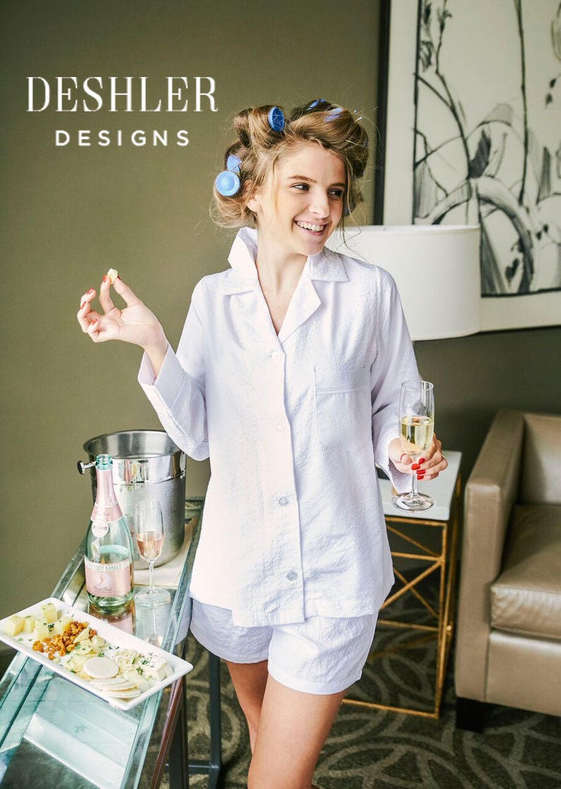 Puede incluir: Una mujer con una camisa de pijama blanca con botones y pantalones cortos a juego. Est&aacute; sosteniendo una copa de champ&aacute;n y un peque&ntilde;o trozo de comida. El texto "DESHLER DESIGNS" es visible en la esquina superior izquierda de la imagen.