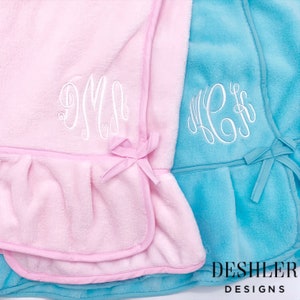 May include: Two plush baby blankets, one pink and one turquoise, with white embroidered monograms. The pink blanket has a pink trim and bow, while the turquoise blanket has a matching trim and bow. The blankets are from Deshler Designs.