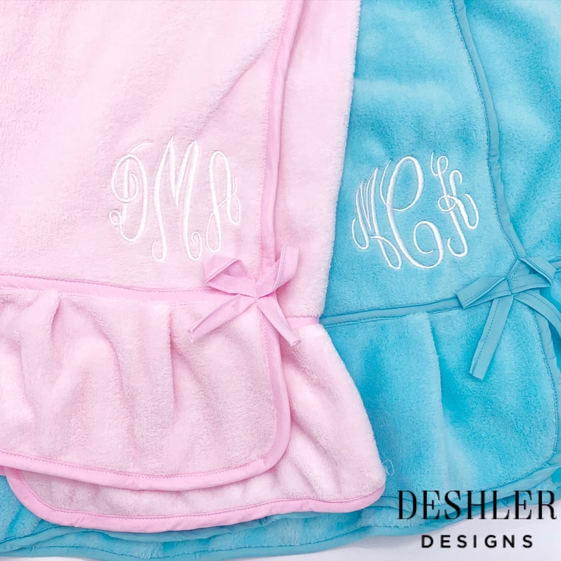 May include: Two plush baby blankets, one pink and one turquoise, with white embroidered monograms. The pink blanket has a pink trim and bow, while the turquoise blanket has a matching trim and bow. The blankets are from Deshler Designs.