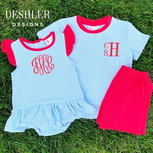 以下が含まれることがあります： 青と白のストライプのシャツと赤いトリムが付いた子供用のお洋服が2着。1着は裾に赤いフリルが付いていて、もう1着は赤いショートパンツ。どちらのお洋服にも赤い刺繍のイニシャルが入っています。