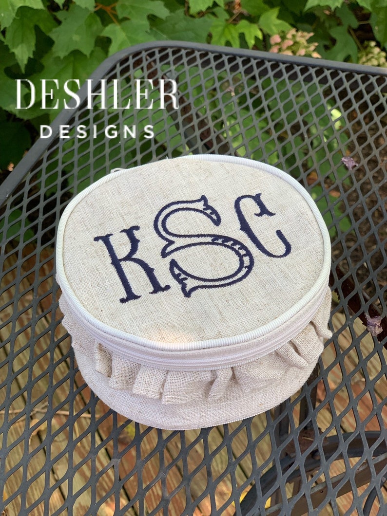 May include: A round, beige linen cosmetic bag with a white zipper and a ruffled edge. The bag is embroidered with the initials "KSC" in navy blue thread.