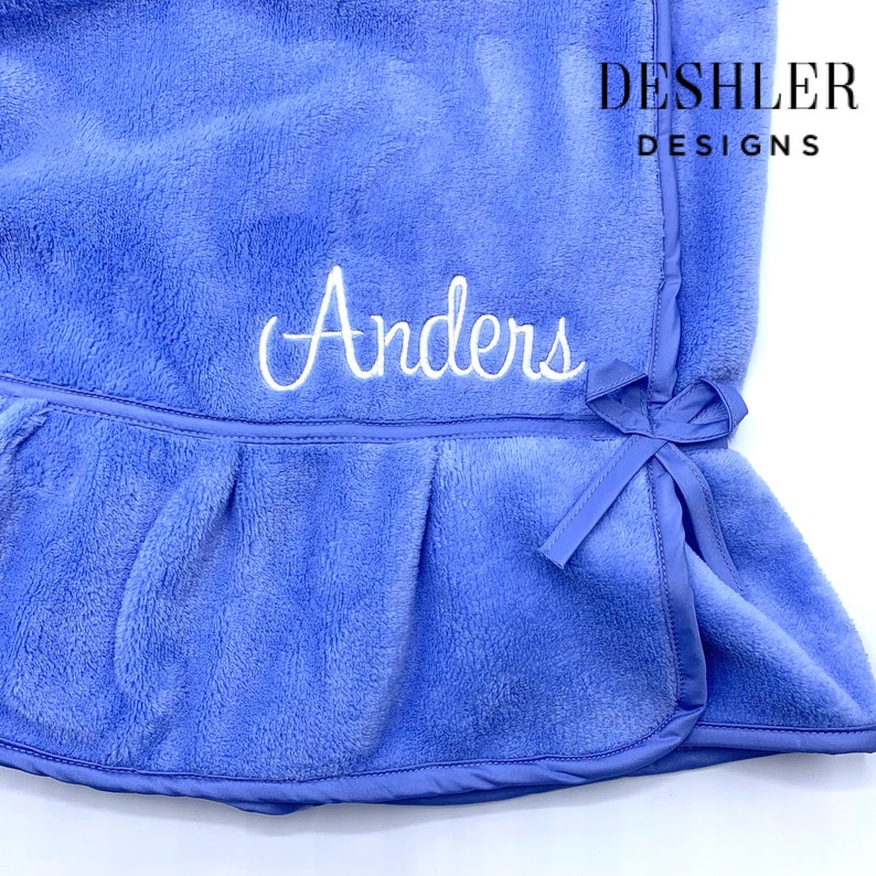 May include: A periwinkle blue baby blanket with the name "Anders" embroidered in white script. The blanket has a ruffled edge and a bow detail. The brand name "Deshler Designs" is printed in black.