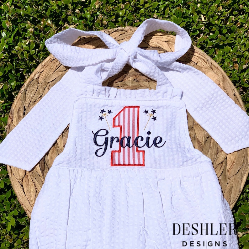 May include: White sleeveless dress with a red and white striped number one and the name Gracie embroidered on the front. The dress has a ruffled neckline and a bow tie at the back.
