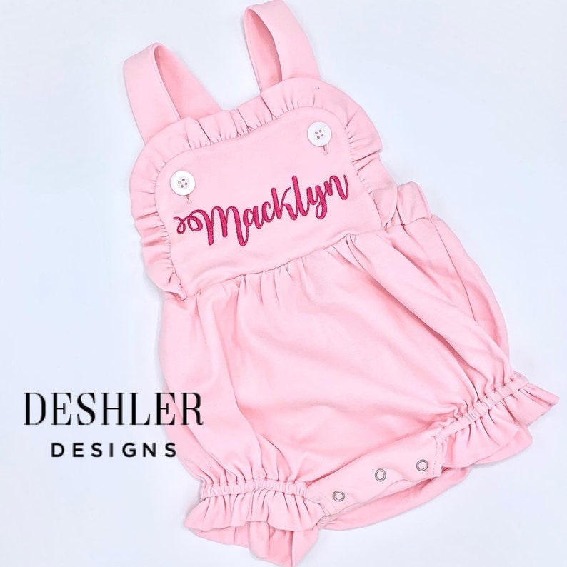 May include: Pink baby romper with ruffled edges and straps. The name "Macklyn" is embroidered in pink script across the front. Features white buttons and snap closures. The brand name "Deshler Designs" is printed in black.