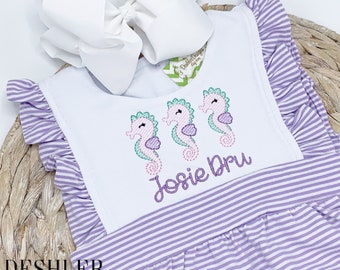 May include: A purple and white striped baby dress with a white bib featuring three embroidered seahorses and the name "Josie Dru".