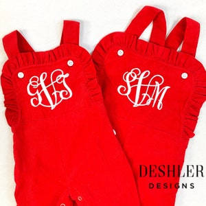 May include: Two red corduroy baby overalls with white ruffled trim and white embroidered monograms. The overalls are on a white background. The text "DESHLER DESIGNS" is visible in the bottom right corner of the image.