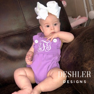 May include: A baby wearing a lavender romper with white embroidered initials and large white bow. The romper has white buttons and the text "DESHLER DESIGNS" is visible.