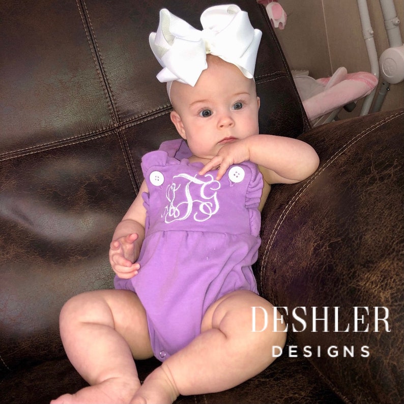 May include: A baby wearing a lavender romper with white embroidered initials and large white bow. The romper has white buttons and the text "DESHLER DESIGNS" is visible.