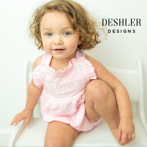 May include: A light pink baby romper with the name "River" embroidered in white. The romper has a ruffled neckline and two white buttons. The image also includes the text "DESHLER DESIGNS".