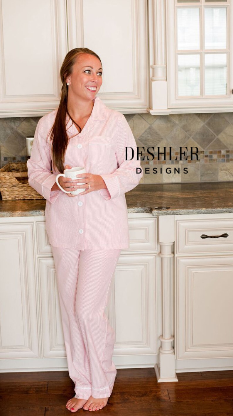 May include: Pink and white striped pajama set with white trim. The long-sleeved top has a button-down front and a breast pocket. The matching pants have a relaxed fit. The pajamas are from Deshler Designs.