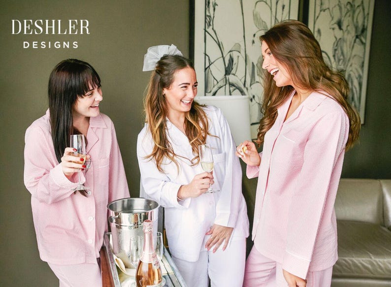Puede incluir: Tres mujeres con pijamas rosas y blancos sonr&iacute;en y sostienen copas de champ&aacute;n. Las mujeres est&aacute;n de pie en una habitaci&oacute;n con una pared gris y una pintura en la pared detr&aacute;s de ellas. El texto "DESHLER DESIGNS" es visible en la esquina superior izquierda de la imagen.