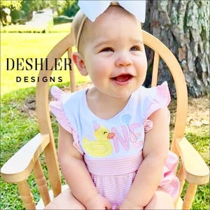 May include: A white and light pink striped romper with ruffled sleeves, featuring an embroidered yellow rubber duck wearing a pink party hat, the word ONE in pink, and a blue balloon. A white bow headband is worn on the head.