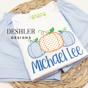 May include: White t-shirt with embroidered pumpkins in orange and blue, with the name "Michael Lee" in blue script. The shirt is paired with blue and white striped shorts. The brand name "Deshler Designs" is visible.